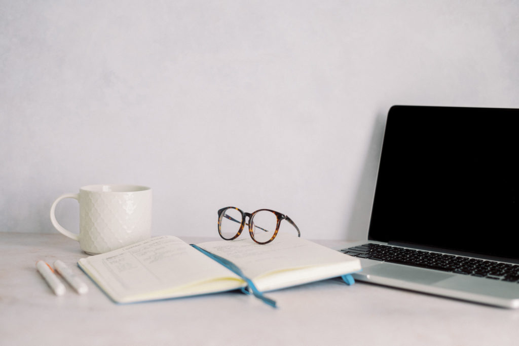 Glasses sitting on top of journal beside laptop and white coffee mug