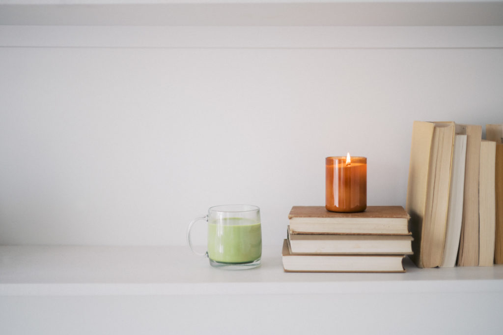 Amber glass candle sitting on stack of 3 books beside a glass mug filled with matcha latte on a bookshelf