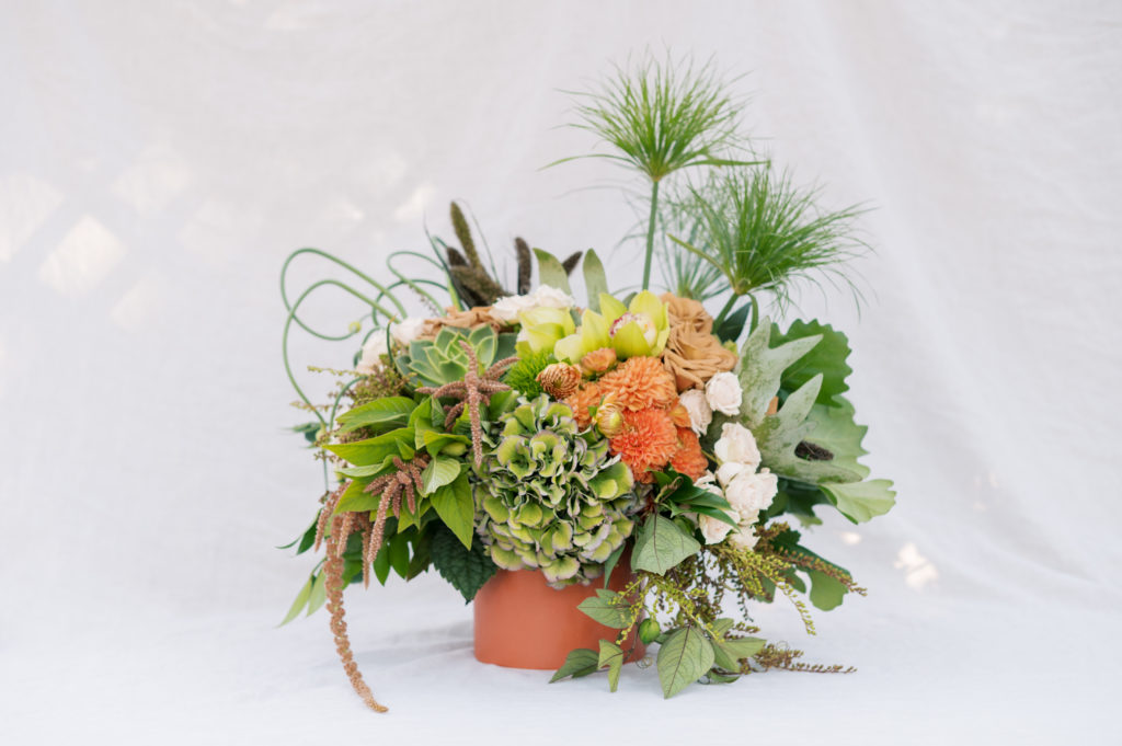 Lush green and orange autumn floral arrangement in terracotta coloured vase designed by Cory Christopher sitting on white linen bedsheet