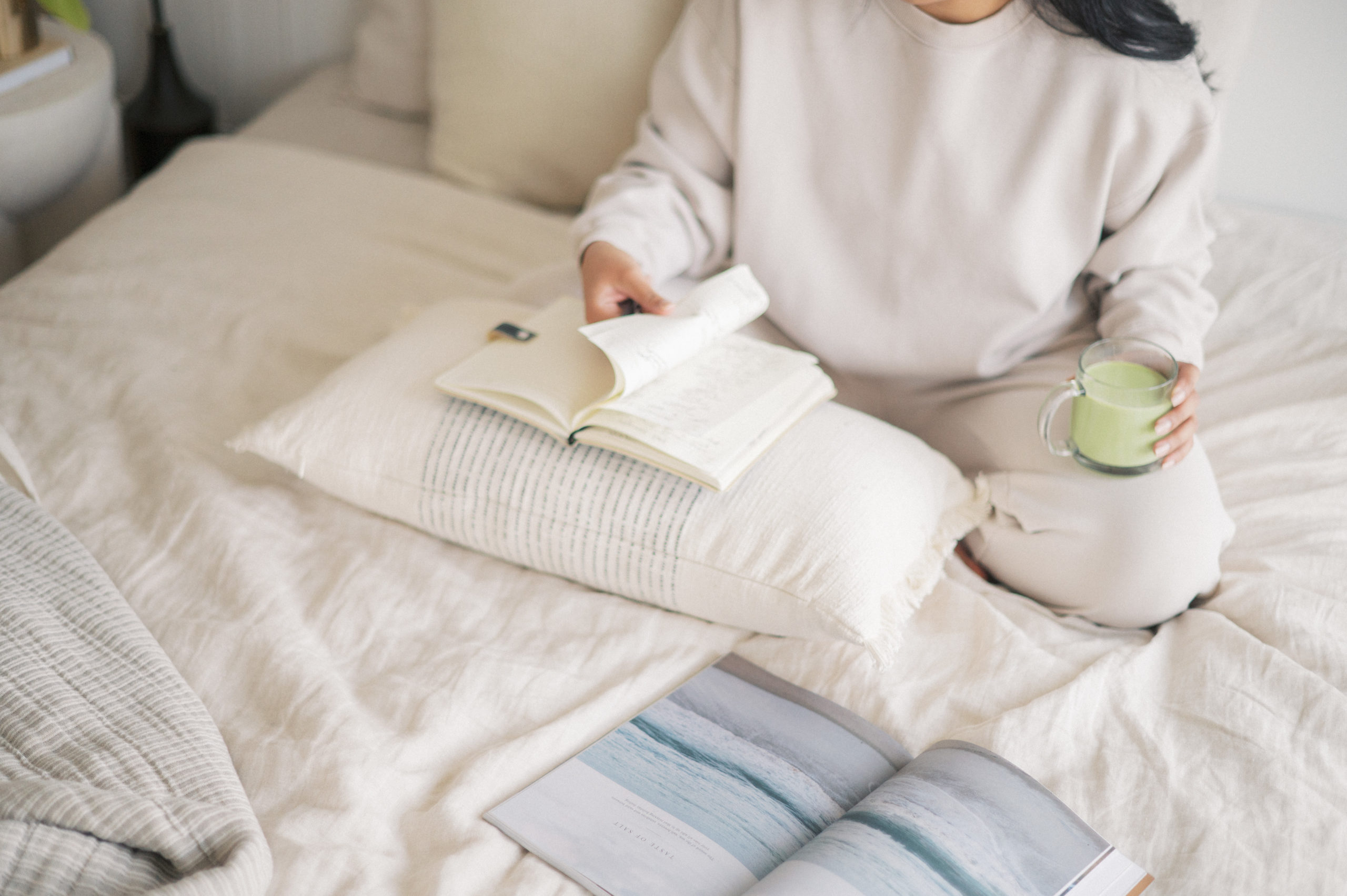 Woman wearing beige sitting on bed with matcha latte flipping through journal