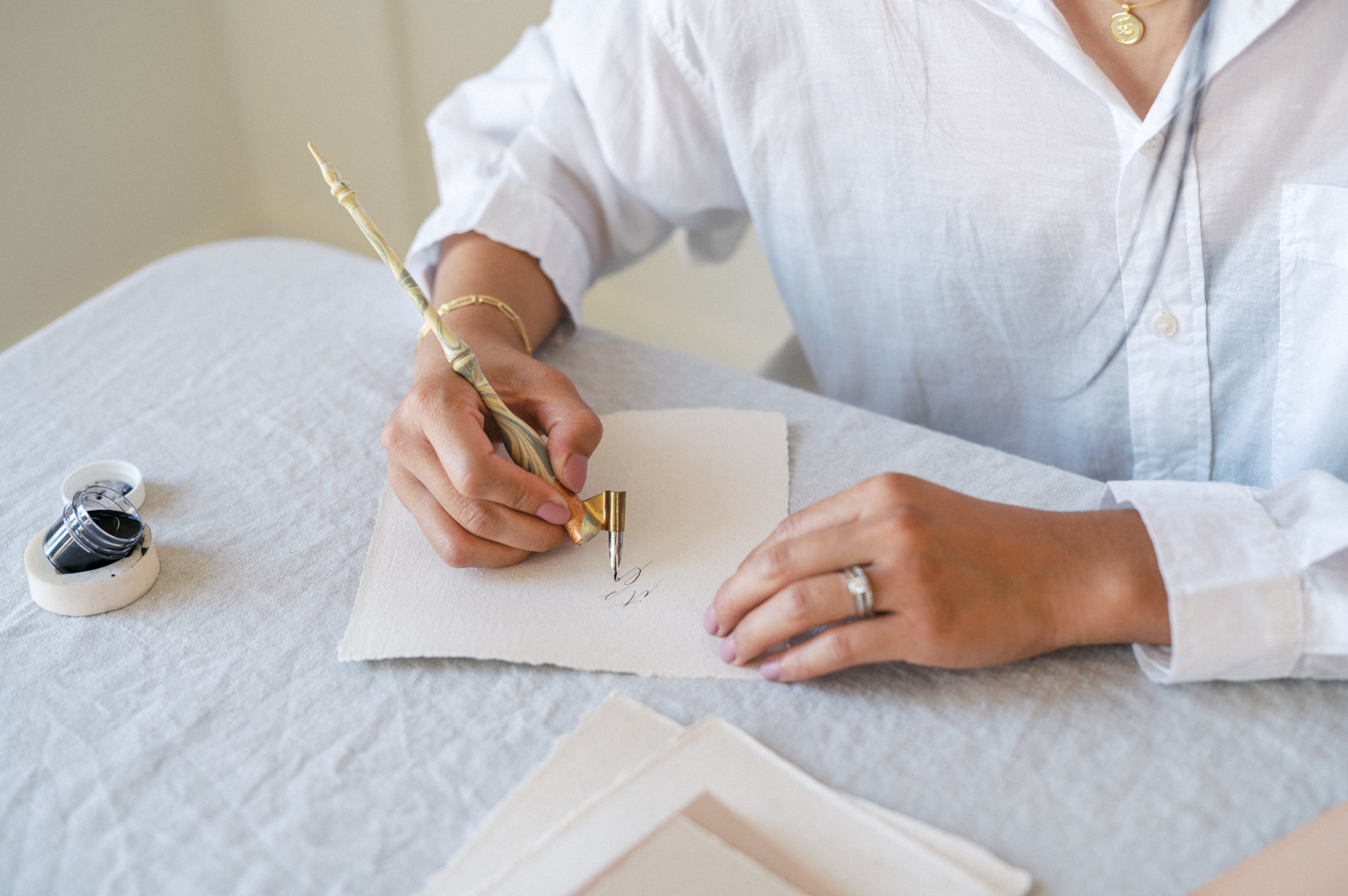 Upclose of womans hands writing calligraphy on neutral coloured paper on a white linen covered table Edmonton Personal Branding Photography 