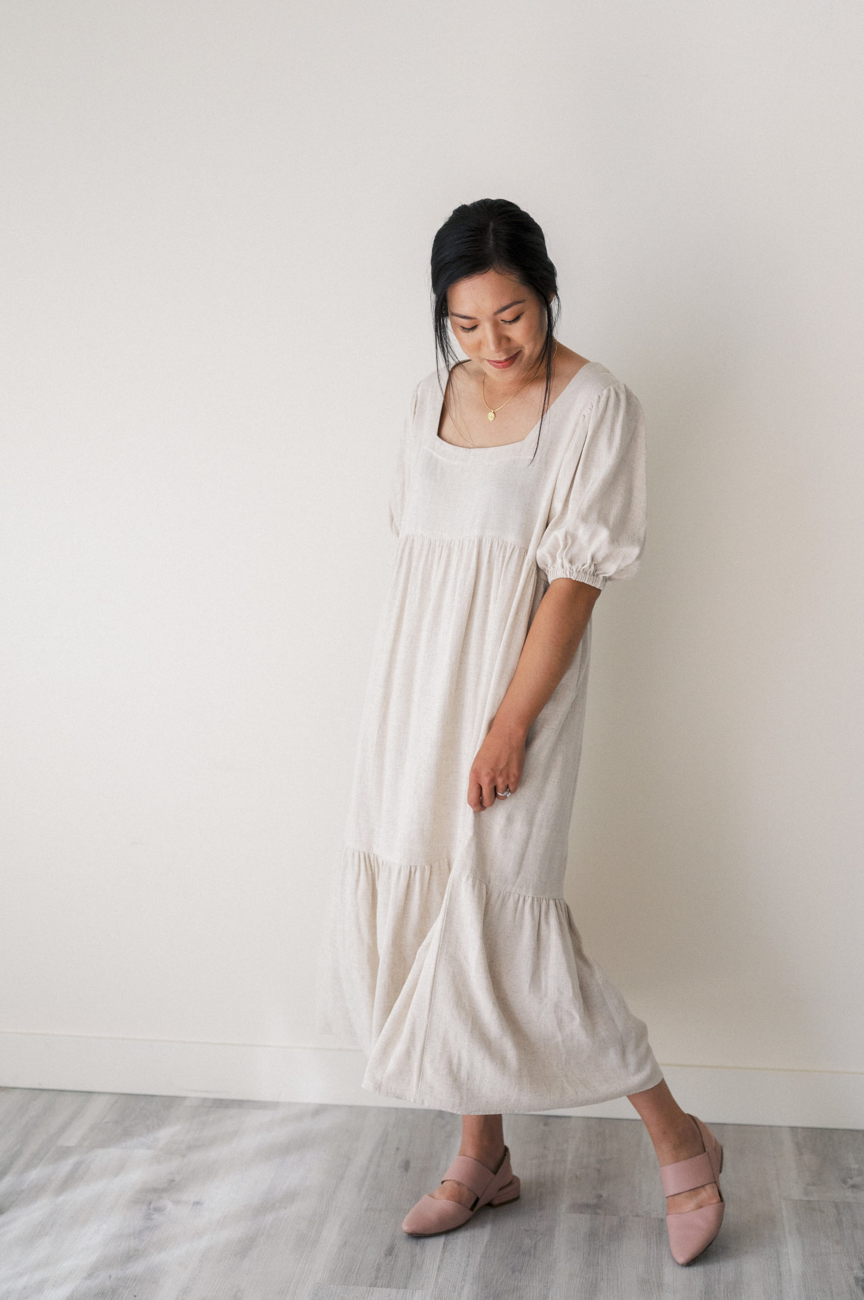 Woman in neutral linen dress swishing dress and looking at the ground 